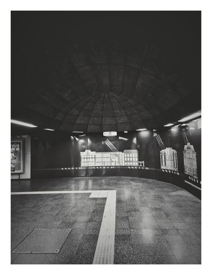Black and white photo of a subway station interior with a curved, domed ceiling. The walls are dark, and a wide, curved section features cutout facades of various museum buildings in a lighter tone, resembling a city skyline. A horizontal white line and a perpendicular white tactile paving strip run across the dark, textured floor in the foreground. A small billboard is visible on the far left wall. The image is oriented vertically.