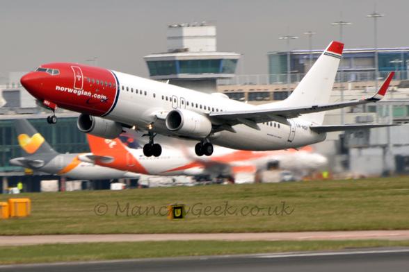 Side view of a twin engined jet airliner flying from right to left at a very low altitude, with undercarriage extended, flaps extended from the rear of the wing, and the nose raised sharply, suggesting it has just taken off.
The plane's forward fuselage is red, with white "Norwegian.com" web address at the bottom.
A thin blue band around the body separated the forward fuselage from the rest of the plane, which is almost entirely white, marred only red up-turned wing-tips, a thick red stripe at the top of the tail, and the black registration "LN-NGV" on the upper rear fuselage.
Green grass fills the bottom of the frame, under the plane, with another couple of airliners, one white and orange, the other white and grey with yellow highlights on the left of the frame, next to a large terminal building in the background.
Jelly from the engine exhaust partially obscures this background.
Grey sky fills the rest of the frame.