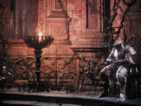 Amnesiac Lapp, identity obscured by heavy armor, rests by a burning brazier before continuing on his parallel journey through the Ringed City. Nominally an ally, restoring his memory summons the troll within.