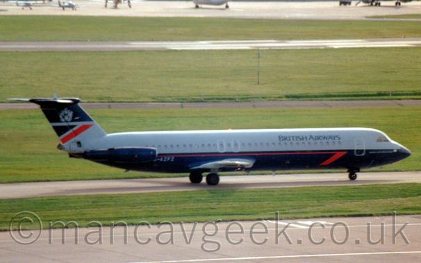 Side view of a twin engined jet airliner with the engines mounted on the sides of the rear fuselage, taxiing from left to right.
The plane is dark grey on the top half of the body, and dark blue of the bottom half, with a red pinstripe just under the passenger cabin windows, ending in half an arrowhead under the forward fuselage.
There are black "British Airways" titles on the upper forward fuselage, and the grey registration "G-AZPZ" just forward of the engine pods on the rear fuselage.
The tail is dark blue at the top and grey at the bottom, with a dark blue triangle at the top rear of the grey part, and a diagonal red stripe below that.
A grey heraldic crest is in the middle of the dark blue part at the top.
Grey concrete apron and green grass fill the foreground, with more grass in the background leading up to a grey runway in the distance.