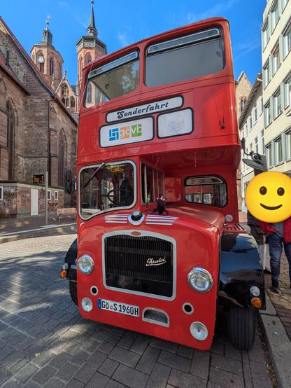 im Vordergrund ein roter Doppeldeckerbus des Typs Bristol Lodekka mit deinen schwarz-roten Einhorn auf der Kühlerhaube, im Hintergrund die Göttinger Johannis Kirche