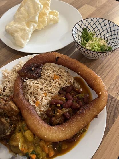 Ein Teller mit nepalesischem Essen. Reis, Gemüse, Fleisch, Soße und Bohnen. Darauf liegt ein Teiggebäck. Dahinter in einer Schale Koreander, Knoblauch und Papadam.