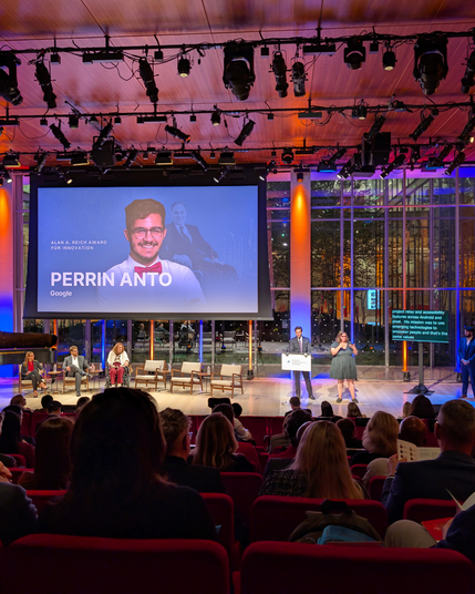 An awards program in a modern auditorium. A large screen announces an innovation award with a headshot. A panel of four is seated on stage. At the podium, a speaker addresses the audience while an ASL interpreter signs nearby. Floor-to-ceiling windows reveal city lights behind the stage.
