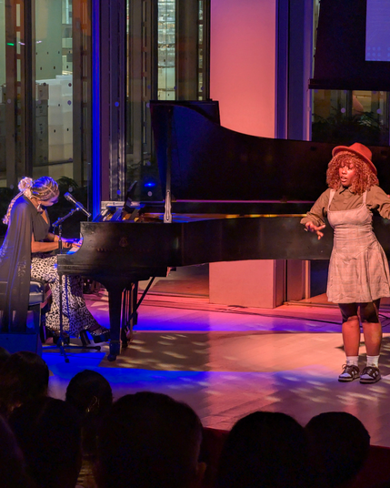A live performance in a glass-walled hall. On the left, a musician in a long black cape-style top plays a grand piano and sings into a mic. On the right, a vocalist in a tan dress and hat gestures expressively. Purple, blue, and amber stage lights wash the scene.