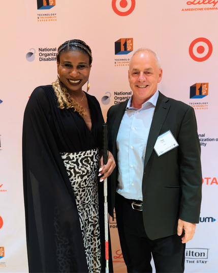 Two people smile on a step-and-repeat with National Organization on Disability logos. The person on the left wears a flowing black top and patterned skirt, has braided hair, and holds a white cane with a red tip. The person on the right wears a dark suit, light shirt, and a name badge.