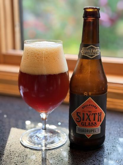 A tall snifter glass full of ruby red ale and a thick bubbly light tan head. next to a brown bottle with a black label that has a red diamond with a blue sash in its center