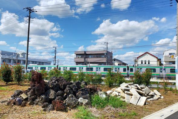 In the background is blue sky with some good weather clouds. Then there are power lines parallel to the horizon and run of the mill buildings. In front of those a JR commuter train is crossing and a couple of its cars made the shot. And then in the foreground is a patch of grass with two piles of stone rubble. The piles are clearly sorted by color, almost black on the left and almost white on the left.
