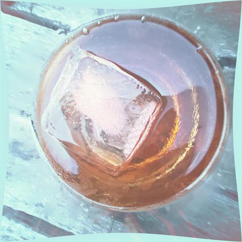 Close-up of a beverage in a glass with a large ice cube, showcasing rich amber colors and condensation on the glass surface. The background is blurred, enhancing the focus on the drink.