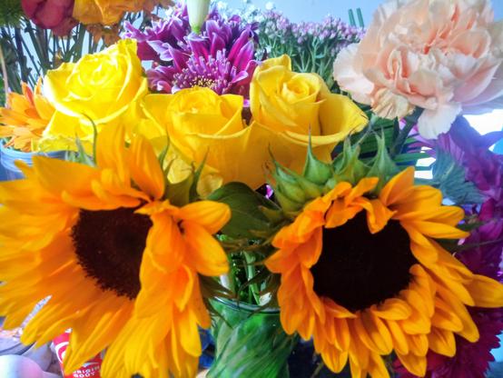A vivid bouquet featuring two large sunflowers with dark centers and golden petals, surrounded by yellow roses, a peach carnation, and purple chrysanthemums. The flowers are fresh and artfully arranged, showcasing rich textures and contrasting colors.