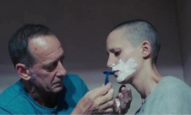 A man is his early 60s is helping shave a woman’s face. She has a buzz cut and is pretending to be his missing son.