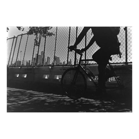A high-contrast black and white photo features the silhouette of a person riding a bicycle in the foreground. The rider is positioned on the right, backlit against a bright sky, making their body and the bike appear completely black. The background includes a chain-link fence on a barrier wall that runs horizontally across the middle of the frame, and a city skyline is visible through the fence in the distance.