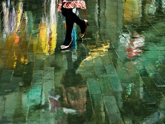 a women wearing gold colored shoes walking on a rainy day, the surounding coloors reflected on the rainy pavement.