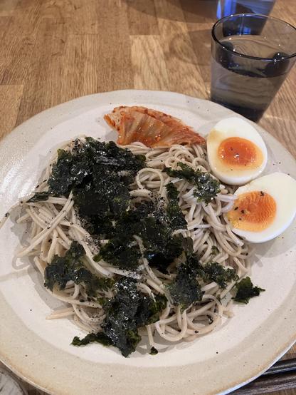 A plate of soba noodles topped with seaweed and black sesame seeds, accompanied by a piece of kimchi and two halved soft-boiled eggs with runny yolks. A glass of water is seen beside the plate, on a wooden table.