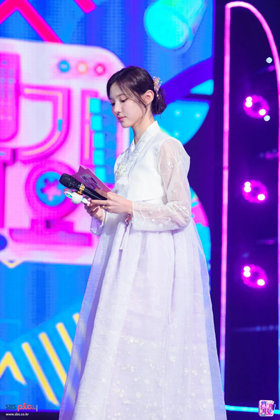 Here's an alt-text description of the image:

A person is standing on a stage, holding a microphone and reading from papers. The person is wearing a long, white dress with long sleeves and a high collar. The background features a blue and purple gradient with abstract shapes, including what appear to be triangular prisms and rounded forms, illuminated with bright lighting. To the left of the person, there's a large, blurred structure with pink and blue elements. In the bottom left corner, the text “sbs play” is visible in white letters on a blue background, and in the bottom right corner, there's a small, square logo with pink and white details.