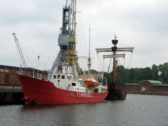 Un bateau-feu à quai dans un port, la coque est rouge et les locaux techniques blancs, le feu est une structure métallique jaune. Derrière se trouve un autre bateau à voiles en bois brun foncé. Sur le quai une grue géante grise impose sa présence. Des bâtiments en briques rouges sont visibles en arrière-plan.
A Lightship in a harbour, the ship is painted in red and white and the light is painted in yellow. Another wooden ship ist visible behind and a big crane is present on the enbankment. In the background are several buildings.