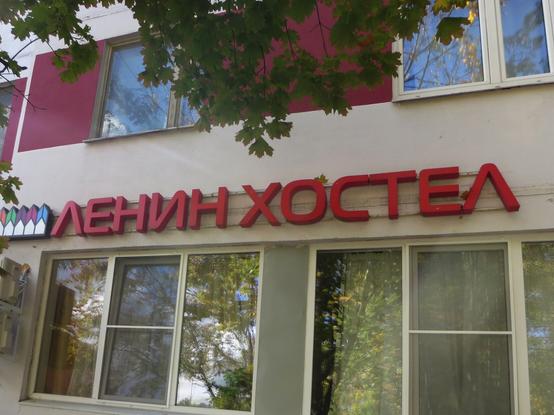 Вывеска «Ленин хостел» над двумя окнами первого этажа многоквартирного дома. Надпись снята снизу вверх, потому что прямо перед вывеской свисают ветки клёна. Конец Ленинского проспекта, Москва. // A “Lenin Hostel” sign hangs above two windows on the first floor of an apartment building. The sign is photographed from the bottom up because maple branches hang directly in front of the sign. End of Leninsky Prospekt, Moscow. // 2025 © unknown designer