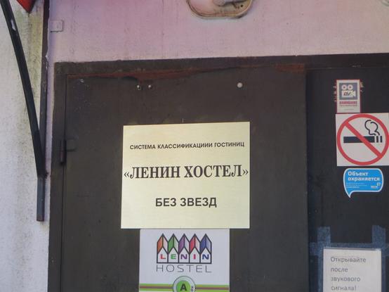 Две вывески на стальной двери: «Система классификации гостиниц. „Ленин хостел“. Без звёзд» и логотип «Lenin hostel». Справа от двери видны несколько стикеров с запретами и предупреждениями и предписание, как нужно открывать входную дверь. // Two signs on a steel door: “Hotel Classification System. Lenin Hostel. No Stars” and the “Lenin Hostel” logotype. To the right of the door are several stickers with restrictions and warnings, along with instructions on how to open the door. // 2025 © unknown designer