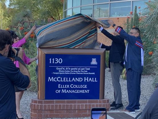 Dark blue sign with white lettering, tribal and university leaders unveiling it by removing a blanket.