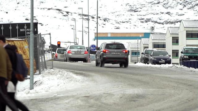 Screenshot aus einer ZDF-Dokumentation über Grönland; vereiste Straße, Berge mit Schnee im Hintergrund, Autoverkehr und eine rote Ampel. Im Hintergrund ein blaues Schild für einen Geh- und Radweg