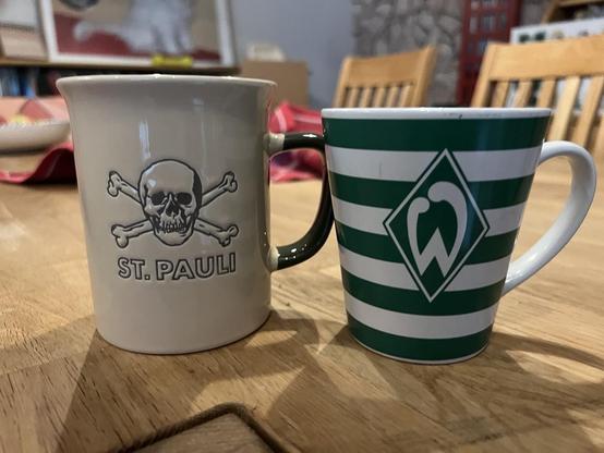 Auf einem Holztisch stehen zwei Tassen. Links eine beige mit einem Totenkopf und der Aufschrift St. Pauli, eine grün-weiß gestreifte Tasse mit der Werder-Raute
