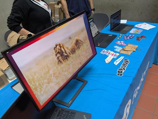 Detailní záběr na stůl stánku s velkým monitorem v popředí a samolepkami v pozadí.