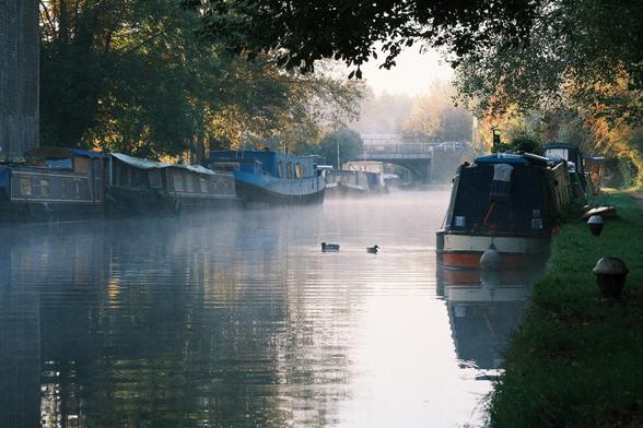 Canal with moored boats on both sides and ducks in the middle. Low fog hangs close to water.