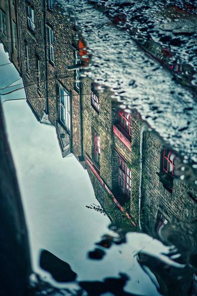 A close-up, low-angle photograph capturing the clear reflection of an old, brick building in a street puddle after the rain. The building is seen upside down in the water, with the red window frames providing a splash of colour against the dark, textured grey of the bricks and the muted, overcast sky. The water's edge creates a dark, irregular border around the reflection.