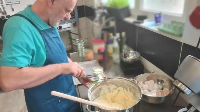 Mann mit Kochschürze steht in einer Profiküche mit viel Edelstahl macht einen Teig, dahinter eine Schüssel mit vielen Eierschalen aufgeschlagener Eier.