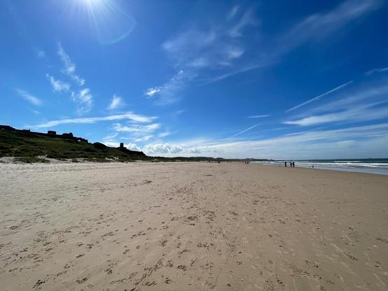 A wide sandy beach in Northumberland stretches out beneath a bright blue sky, with gentle waves rolling in from the North Sea. Sparse groups of people walk along the shoreline, leaving scattered footprints across the smooth sand. Low grassy dunes rise on the left, with dark silhouettes of old stone structures and buildings along the ridge. Wispy clouds and streaked contrails cut across the sky, while the sun sits high, casting a soft glare. The beach appears open and expansive, with the coastline extending far into the distance.