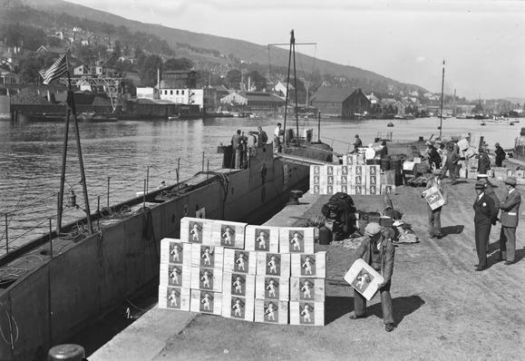 A black and white photo of a 1910s submarine, the Nautilus, docked up in Bergen, being stocked with chocolate.