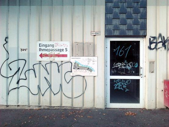 Tür in der Ruine des Ihmezentrums in Hannover-Linden. Der Beton um die Tür wurde sichtbar frisch, aber auch etwas eilig übermalt, vermutlich, um Graffiti zu verdecken. Die ersten Graffiti sind schon wieder da. Rechts neben der Tür ein Wegweiser zum Eingang in das Haus Ihmepassage 5 und ein Lageplan des Ihmezentrums.