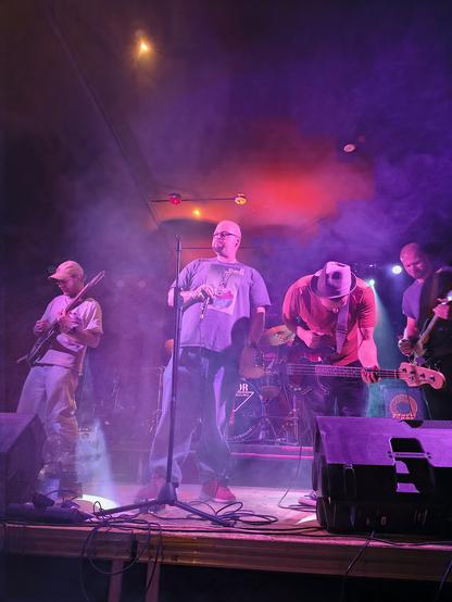Auf einer un violettes Licht und Nebel gefüllten Bühne sind in leichter Untersicht zwei Gitarristen, ein Bassist und ein Sänger zu sehen