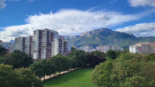 Premier plan : parc de la Villeneuve, une pelouse bordée d'arbres, un chemin de terre qui va vers une petite colline d'herbe
Deuxième plan : la galerie de l'arlequin, barres d'immeubles colorés et situant
Troisième plan au-dessus des immeubles : le Vercors et notamment le Moucherotte, montagne toute arrondie et recouverte d'arbres.
Au-dessus du Moucherotte, ciel bleu et nuages blancs