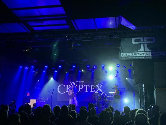 The Cryptex performing on stage. On the left, the singer at a piano, in the back, a masked guitar player, on the right the drummer. Lighting is blueish. A big banner in the back with “The Cryptex” projected on it.