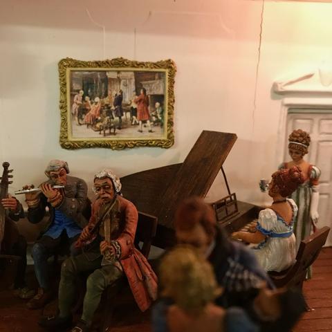 Miniature diorama depicting an elegant 18th-century music salon with tiny figurines playing instruments, including a violin, flute, and piano, while others listen and converse. A framed painting hangs on the wall above the musicians, and the scene is warmly lit.
