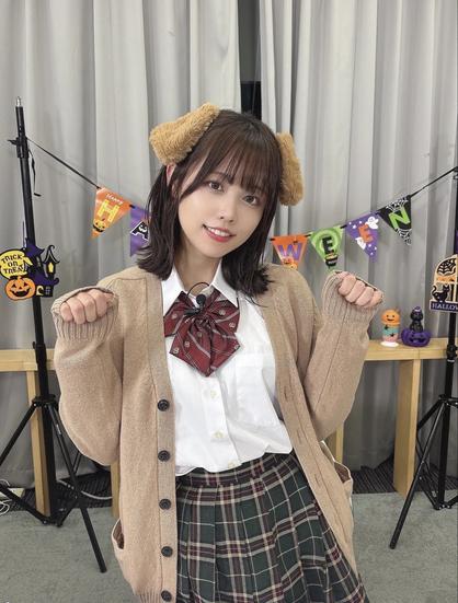 A young woman is posing indoors wearing a school uniform with a plaid skirt, white shirt, red ribbon tie, and beige cardigan. She has fluffy brown animal ear headbands and is making a playful pose with her hands like paws. Behind her, there are Halloween decorations including a banner, pumpkins, and themed props.