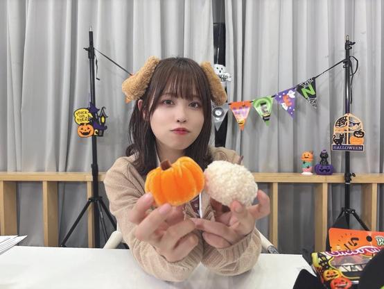 A young woman is sitting at a table wearing a beige cardigan and fluffy brown animal ear headband. She is holding two small decorative pumpkins, one orange and one white, toward the camera. Behind her, Halloween decorations including a festive banner, pumpkins, and themed ornaments are displayed.