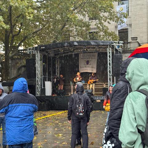 Die Musik Spiel auf der Bühne, aber davor regnet es in Strömen. Weniger Leute (die waren unter die Bäume geflohen), mehr Regenschirme. Im Hintergrund der Bühne hängt ein Banner: "Wir können alles. Außer Sicherheit."