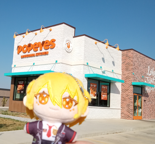 Photo of Don Quixote the plush from Limbus Company in front of a Popeye's, granted with some distance as the structure looks blurry and far away. She's looking at us.