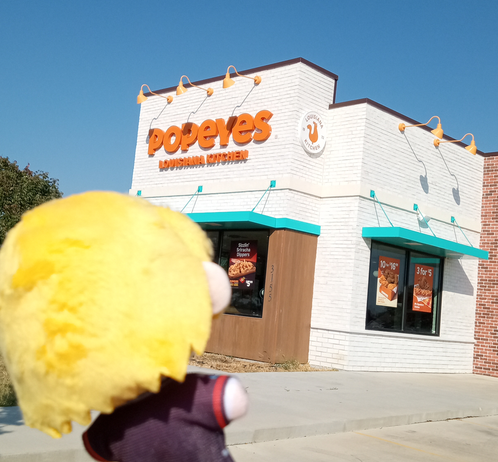 Photo of Don Quixote the plush from Limbus Company in front of a Popeye's, granted with some distance as the structure looks blurry and far away. She's looking at the building now.