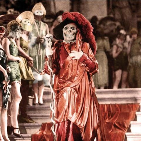 A figure wearing a skeleton mask and a red attire stands on the steps of the Opera House during a masked bal.