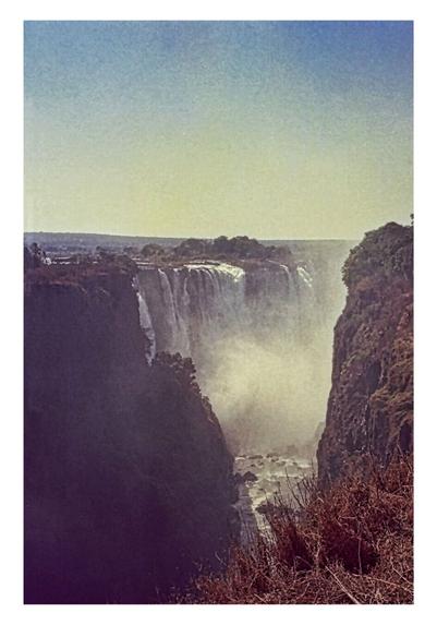 Majestic waterfall cascades between lush cliffs, mist rising into the clear sky. Rocky foreground frames the tranquil yet powerful scene.