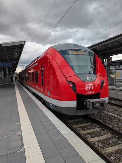 Ein roter Zug von der Deutschen Bahn, Baureihe 1440 von vorne fotografiert. Recht rundlicher und windschnitter Zug. Links davon Bahnsteig. Bewölkter Himmel mit einsetzenden Sonnenuntergang im Hintergrund.