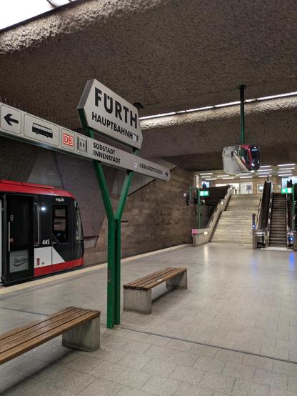 U-Bahn Station Fürth Hauptbahnhof. Der Bahnsteig ist grau gepflastert, wo zwei Holzbänke stehen. Außerdem grünes Stationsschild. Die Wände gemauert und zum Teil mit Waschbeton ausgekleidet. 80er Jahre Stil. Links sieht man einen kleinen Teil eines rot-schwarz-weißen U-Bahn Zuges. Im Hintergrund Treppenaufgänge.