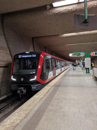 Nochmal eine U-Bahn Station, diesmal mit Fokus auf die U-Bahn. Ein rot-schwarz-weißer Zug mit der Wagennummer 533, der links vom Bahnsteig steht. Das Ziel ist U1 Langwasser Süd. Der Zug wirkt sehr modern. Auf dem Bahnsteig sieht man einige Menschen, außerdem Zugzielanzeigen und wieder die Waschbetondecke. Schummerig beleuchtet.