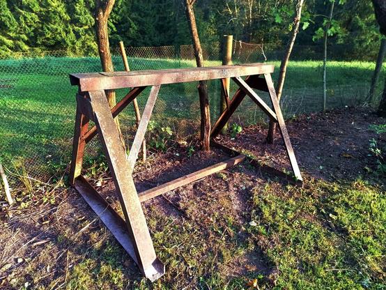 A large steel sawhorse, about 1.2m tall and 2.5m wide. It's rusty and made of thick C-channels with diagonals and a bottom link. It's now sitting in a recently cleared part of the garden by a fence.