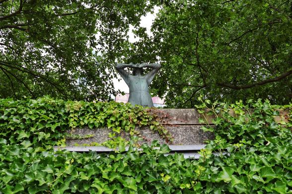 A view from below of the bronze statue ‘Der Rufer’ in Bremen, depicting a human figure with hands raised to their mouth as if calling out. The statue is partially framed by lush green ivy and dense foliage, creating a natural contrast with the bronze figure and the stone wall in front of it.