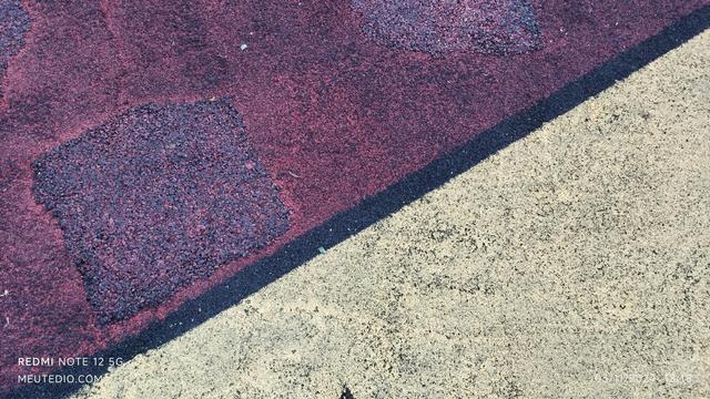 Uma imagem de close-up de uma superfície com textura dividida em duas seções diagonais. A seção superior esquerda é vermelha e granulada, enquanto a seção inferior direita é amarela e também granulada. A divisão entre as duas seções é diagonal, atravessando a imagem do canto superior direito ao canto inferior esquerdo. Pequenos detritos, como galhos finos ou folhas, estão espalhados em ambas as seções.