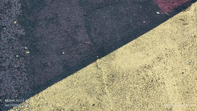 Uma imagem de close-up de uma superfície com textura dividida em duas seções diagonais. A seção superior esquerda é preta e granulada, enquanto a seção inferior direita é amarela e também granulada. A divisão entre as duas seções é diagonal, atravessando a imagem do canto superior direito ao canto inferior esquerdo. Pequenos detritos, como galhos finos ou folhas, estão espalhados em ambas as seções.