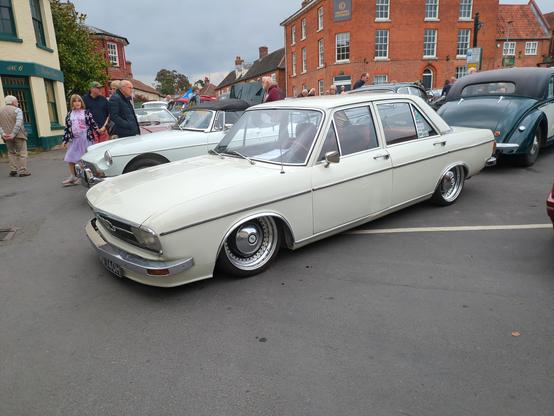 Lowered so much the bodywork is practically scraping the ground, a white Audi 100 saloon sits among more traditonal classic cars in Reepham Market Place with, in the background, a street of 18th century disappearing into the distance. The Audi has deep-dished wheels with chrome hubcaps, but almost the entire top half of the wheel is hidden behind the wheel arch. Several people are admiring other cars in the background.